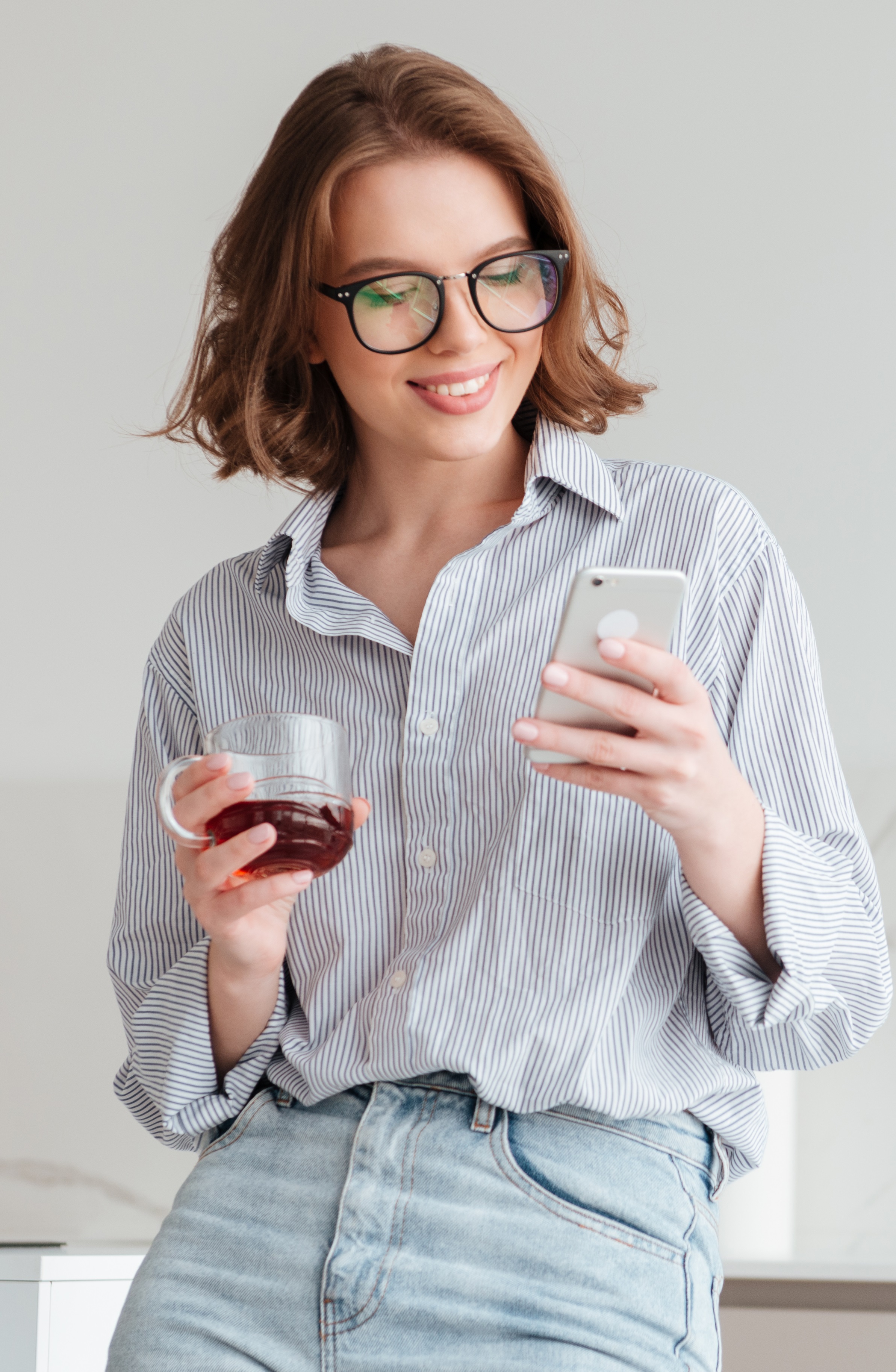 Portrait of a happy attractive woman using mobile phone while holding cup of tea and leaning on a table with laptop computer at home